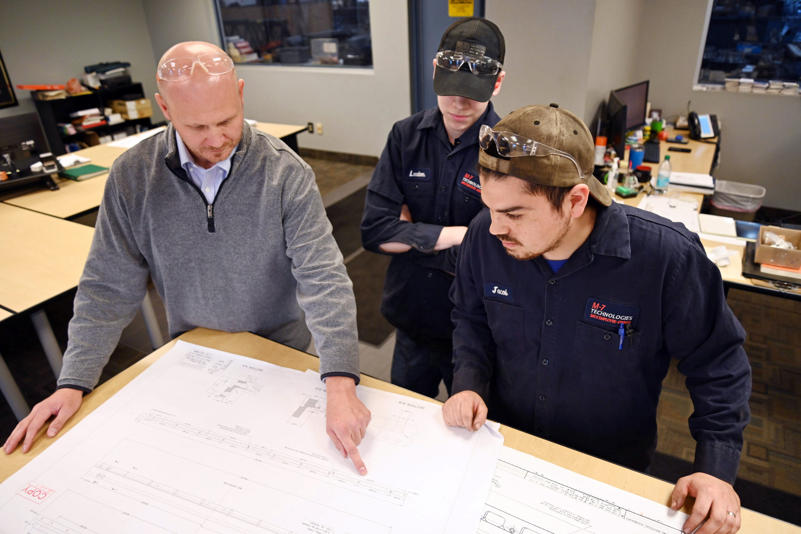Three guys looking at blue prints on a drawing table, guy on the left is pointing at the paper